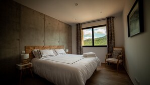 Ein Schlafzimmer mit einem großen Bett, einem Nachttisch, einem Sessel und einem Fenster mit Blick auf Grünflächen.