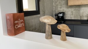 Eine Küchenarbeitsplatte mit einer Kaffeemaschine, einem Buch mit dem Titel 'Le Meilleur des Chefs' und zwei Holz-Pilz-Skulpturen.