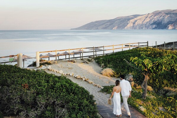 Un paseo marítimo con valla de madera y sombrillas de paja, que lleva a una playa con vista al mar y montañas distantes.