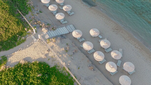 An aerial view of a beach with rows of sun loungers and umbrellas, a wooden walkway, and clear turquoise water.