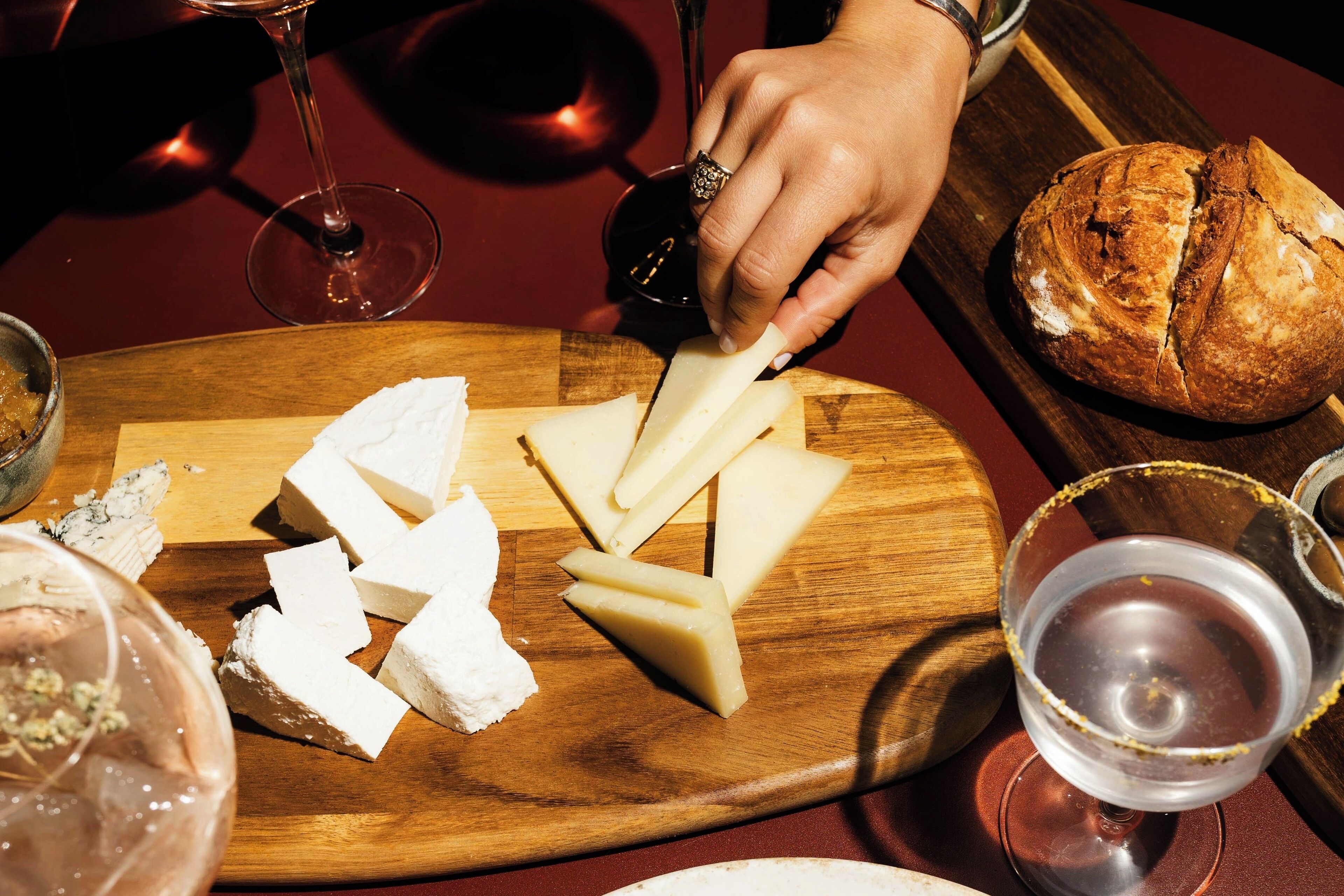 A wooden cutting board with cheese and a knife, a glass of red wine, and a loaf of bread on a table.