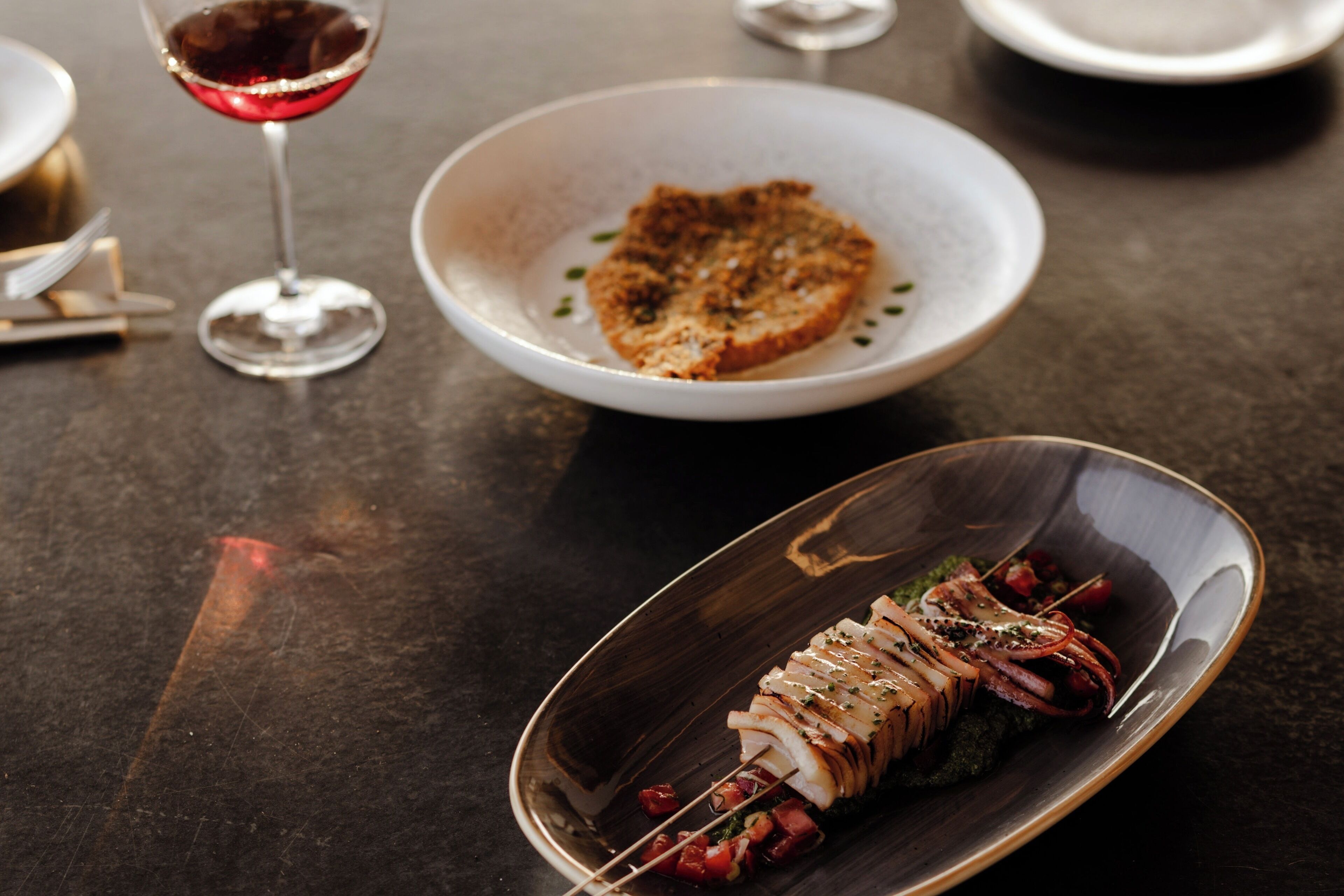 A dining table with a plate of grilled fish, a bowl of bread, and a glass of red wine.