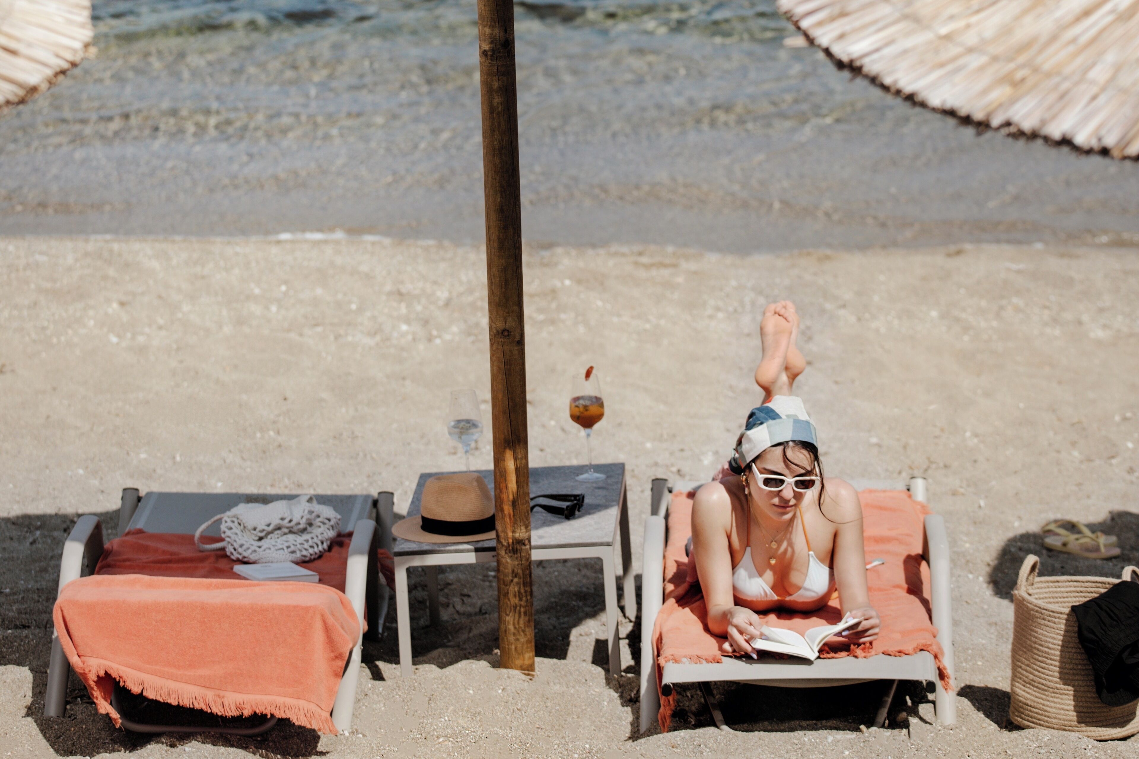 A beach scene with two lounge chairs, one occupied by a person reading a book, and another with a hat and a bag.