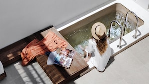 A bathtub with a person sitting inside, a wooden bench with a magazine, and a towel on the floor.
