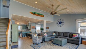 A spacious living area with a large gray sectional sofa, a patterned rug, and a ceiling fan. A kitchen is visible in the background, and a compass wall decoration is present. - Enjoy this oceanfront 3-bedroom Sea Rie-Treat, in North Topsail Beach. (North Topsail Beach)