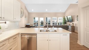 A modern kitchen with white cabinetry, a dishwasher, and a large island with a sink. There is a clock on the wall and a television mounted above the island. - Dark Side Of The Dune - 4 Bedroom, Sleeps 14 - Gulf View in gated community (Port Bolivar)