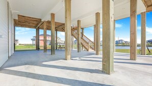 An open interior space with concrete flooring and wooden beams, large windows offering views of a lake and buildings. - Dark Side Of The Dune - 4 Bedroom, Sleeps 14 - Gulf View in gated community (Port Bolivar)