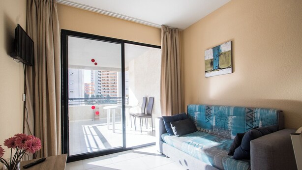 Ein modernes Wohnzimmer mit einer Couch, einem Fernseher, einer Blumenvase und einem Balkon mit Blick auf Gebäude.