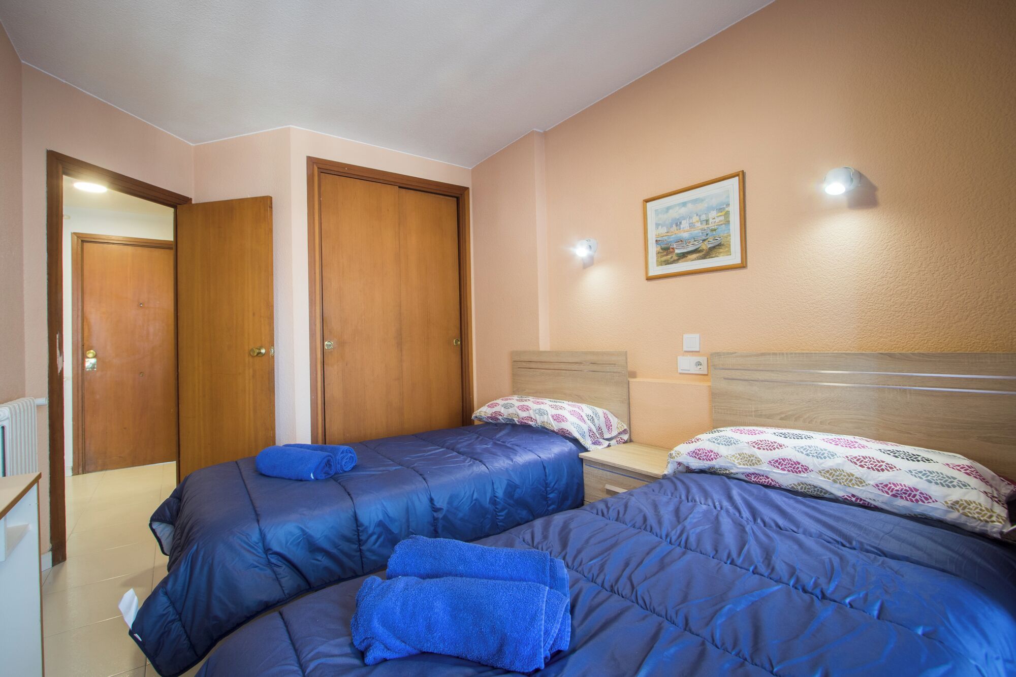A bedroom with two beds, a wooden wardrobe, a painting on the wall, and a nightstand with a lamp.