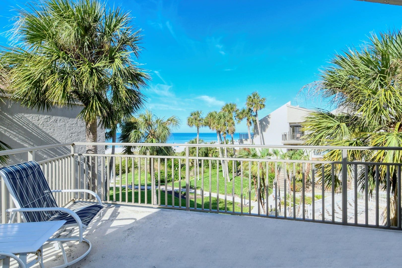 A balcony with a view of the ocean, palm trees, and a white building.