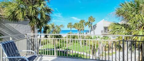 A balcony with a view of the ocean, palm trees, and a white building.