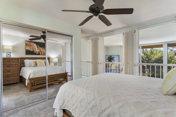 A bedroom with a large bed, a ceiling fan, a dresser, and a view of the outdoors.