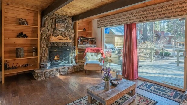 A cozy cabin interior with a stone fireplace, wooden shelves, a comfortable chair, and a large sliding glass door to a deck.