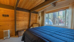 A wooden cabin room with a bed, a radiator, a window with curtains, and a chair.