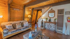 A cozy cabin interior with a sofa, a coffee table, a fireplace, and a staircase leading to another level.