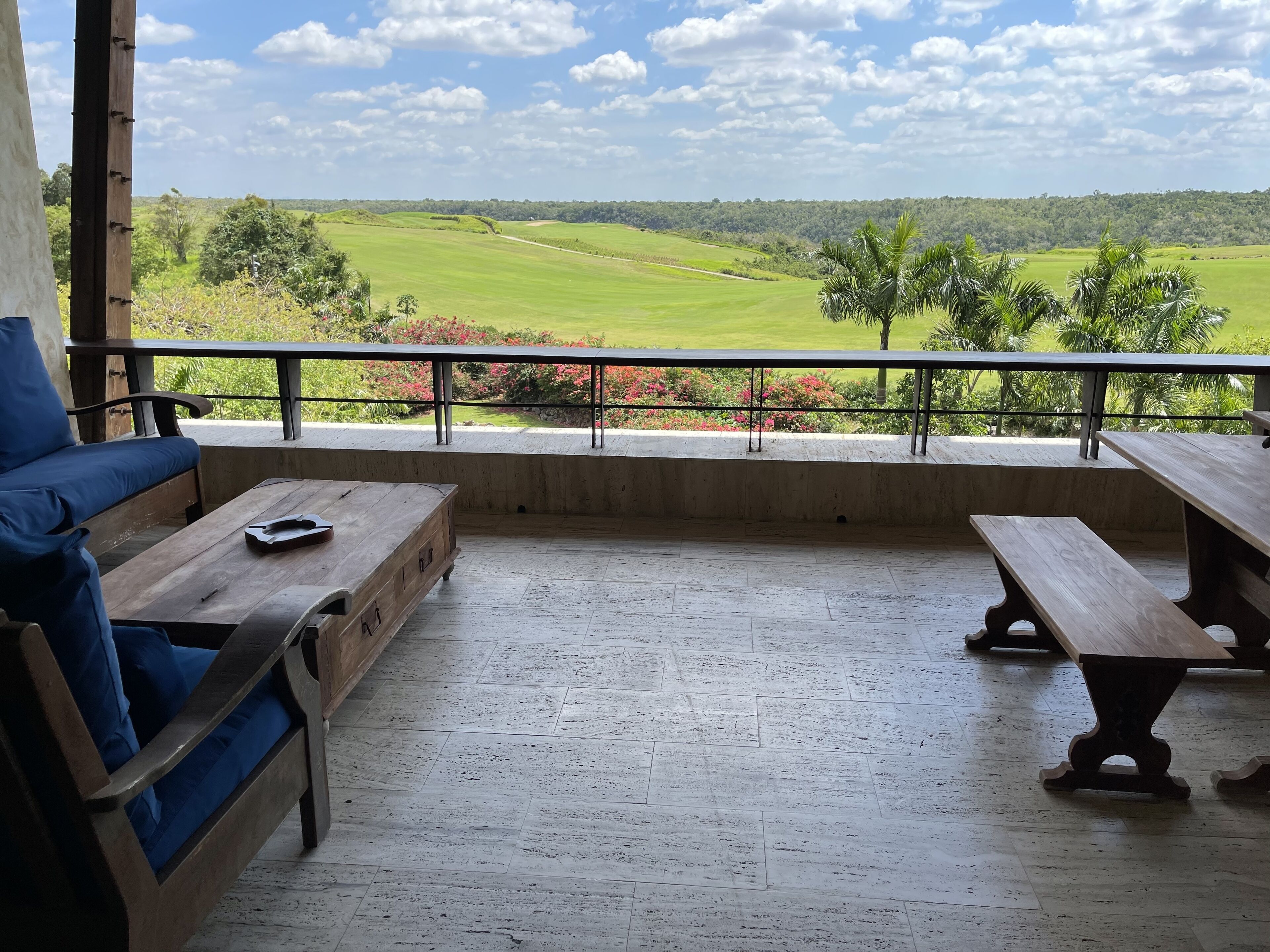 A balcony with a wooden bench and a sofa, overlooking a green landscape with trees and a clear sky.