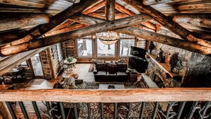 Interior rústico de cabana, com teto alto, vigas de madeira, lareira aconchegante e uma grande janela com vista para uma paisagem nevada.