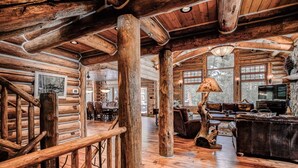 Interior espaçoso de uma cabana de toras, com uma escada de madeira, uma grande mesa de jantar rústica e um espaço de estar acolhedor.