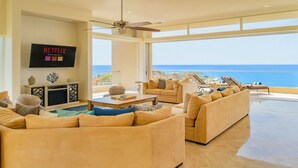 A spacious living room with a large sofa set, a coffee table, and a flat-screen TV mounted on the wall. The room has large windows offering a view of the ocean. - Villa Del Mar-5 Star Luxury Villa with Epic Views (Cabo San Lucas)