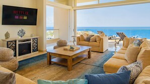 A spacious living room with a large window overlooking the sea, a fireplace, and a flat-screen TV mounted on the wall. - Villa Del Mar-5 Star Luxury Villa with Epic Views (Cabo San Lucas)