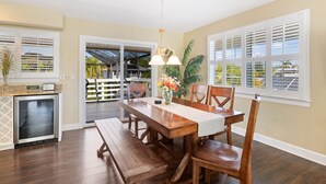 A dining area with a wooden table, benches, and chairs. There is a refrigerator, a plant, and a window with shutters.