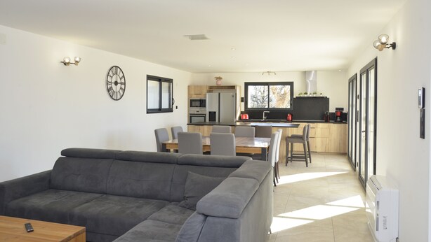 Un salon moderne avec un canapé gris, une table à manger avec des chaises, une cuisine en arrière-plan et une grande fenêtre laissant entrer la lumière naturelle.