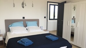 Un lit bien fait, avec deux oreillers, une couverture bleu foncé et deux serviettes pliées. On y trouve également une armoire avec un miroir et une fenêtre équipée de stores.