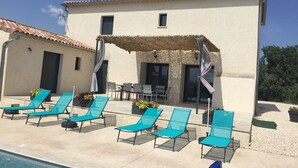 Un espace aménagé au bord de la piscine, avec des chaises longues, et un bâtiment doté d’un toit recouvert de tuiles.
