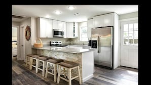 A modern kitchen with white cabinets, a stainless steel refrigerator, a microwave, and a granite countertop. There are three wooden stools and a wicker basket on the counter. - Discover Anchors Down, a charming 3-bedroom duplex in Oak Island, perfect for family getaways! (Oak Island)