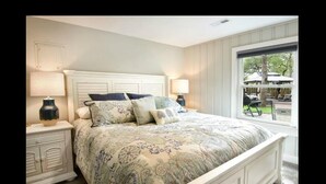 A bedroom with a large bed, two bedside tables with lamps, a window with a view of the outdoors, and a door. - Discover Anchors Down, a charming 3-bedroom duplex in Oak Island, perfect for family getaways! (Oak Island)