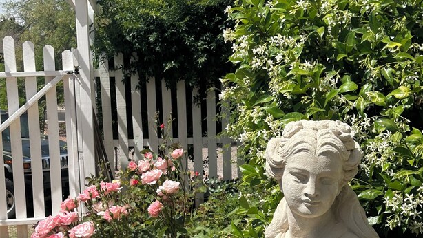 A garden statue holding a flower, surrounded by blooming pink roses and lush greenery. - Historic Schoolhouse Flat by Verde Canyon Railroad (Clarkdale)