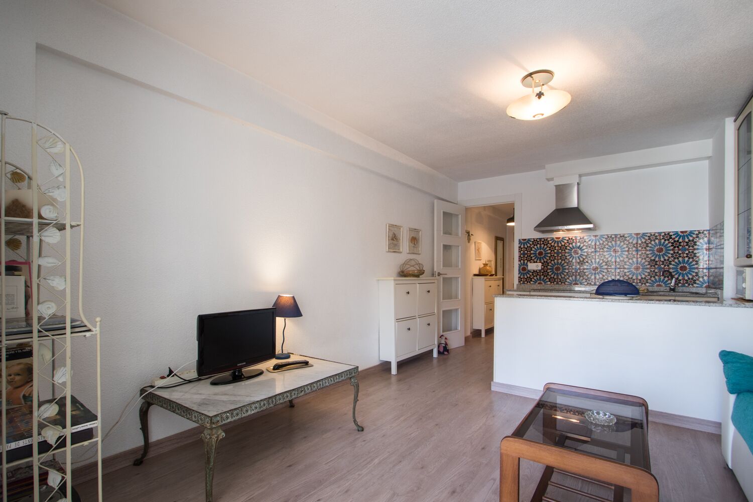 Een ruimtelijk ingerichte leefruimte met een televisie, een lamp, een klein tafeltje en een keukenhoek met een kleurrijke achterwand.