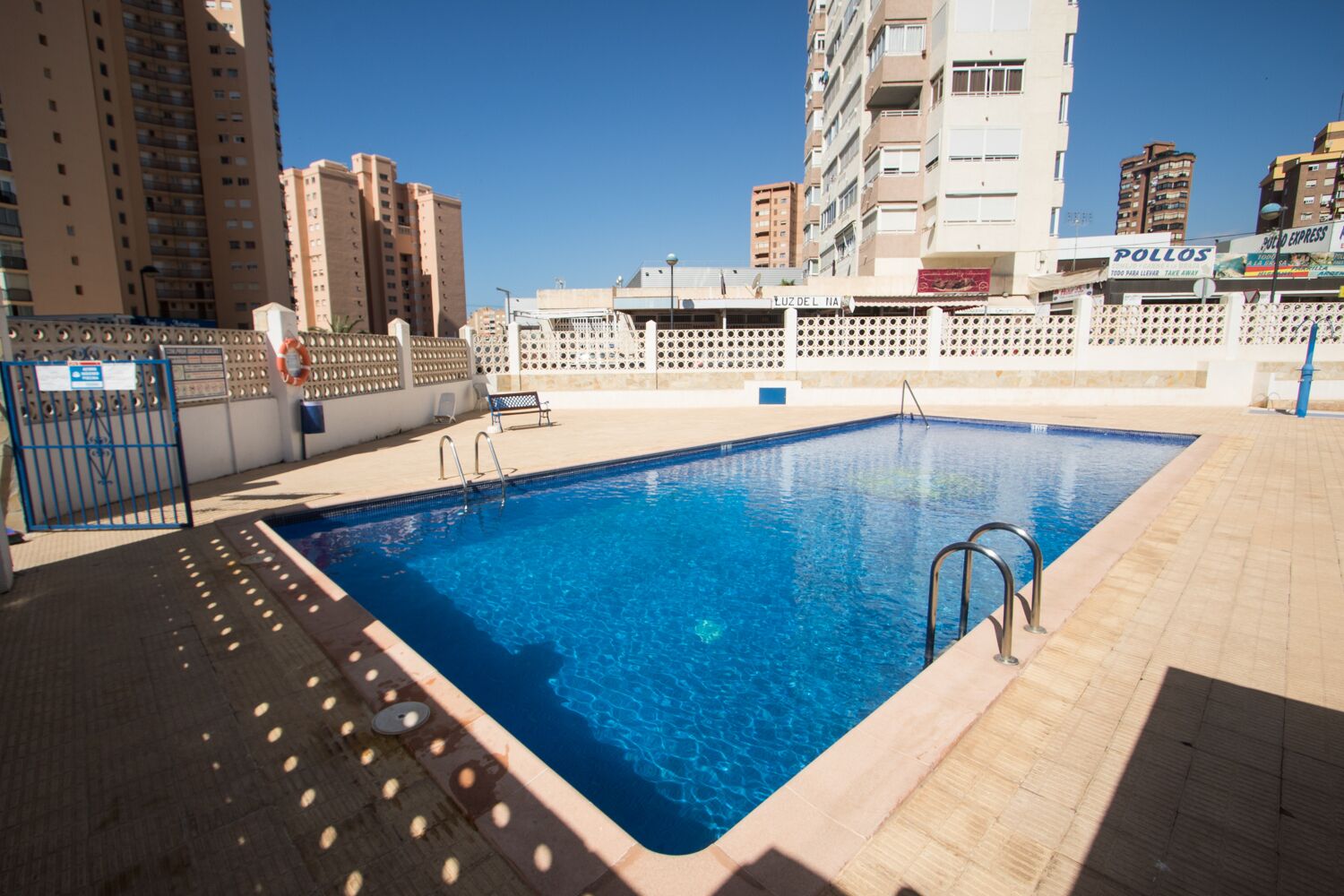 Piscina rectangular con escaleras metálicas en un lateral, rodeada por una solería empavedada.
