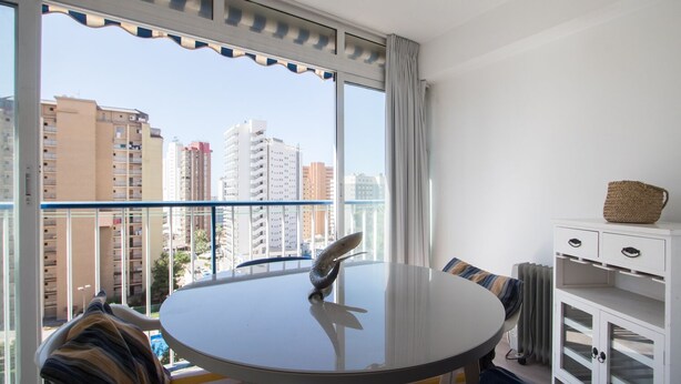 Een balkon met een ronde tafel en stoelen, een wit kastje met lades en uitzicht op hoge gebouwen.