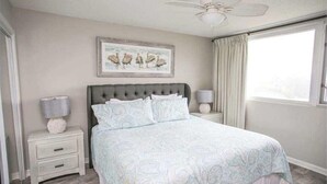 A bedroom with a large bed, two bedside tables with lamps, a ceiling fan, and a window with curtains. - Fun & Easy Access to Everything! Islander 216 (Destin)