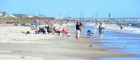 Une plage où l’on voit des gens pêcher et s’asseoir sur des chaises, une jetée en arrière-plan et un ciel dégagé.