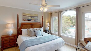 A bedroom with a large bed, a ceiling fan, a window with curtains, a nightstand with a lamp, and a chair. - Brand New Listing! Heated Pool Access, Steps from Beach, Restaurants & Shopping! (Tybee Island)