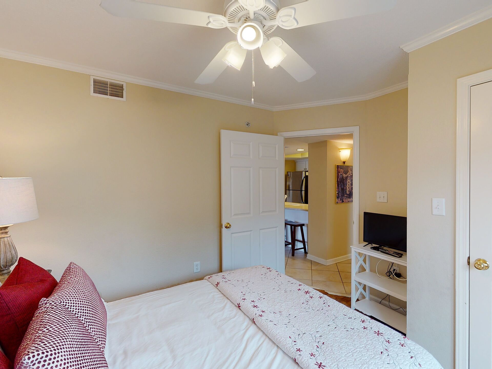 A bedroom with a bed, a ceiling fan, a door leading to a kitchen, and a TV stand.