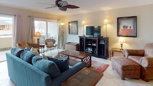 A living room with a sofa, armchair, coffee table, TV stand, and ceiling fan. - Brand New Listing! Heated Pool Access, Steps from Beach, Restaurants & Shopping! (Tybee Island)
