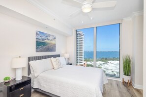 A bedroom with a bed, nightstands, a ceiling fan, a large window with a view of a building and ocean, and a potted plant.