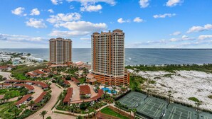 Um resort costeiro com dois edifícios altos, uma praia e uma quadra de tênis.