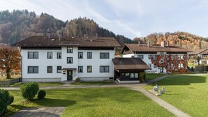 Um edifício de vários andares com telhado em telhas, cercado por um gramado bem cuidado e uma encosta arborizada ao fundo.