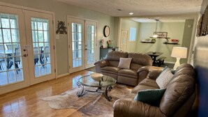 A spacious living room with a leather sofa, glass coffee table, and a large rug. There are glass doors leading to an outdoor area with chairs. - Cocktail Cove 4 bedroom 2 bath by Jamestown Marina (Jamestown)