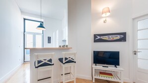 A modern living room with a flat-screen TV, two white chairs, a small table, a wall-mounted lamp, and a fish artwork.
