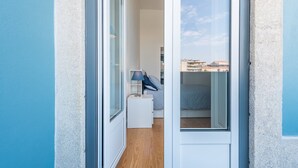 A narrow hallway leading to a bedroom with a bed, a nightstand, and a lamp. A window reveals a cityscape view.