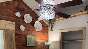 Une chambre avec un mur en bois, un ventilateur de plafond, une cheminée et un lit recouvert d’une couvre-lit à motifs.
