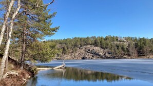 A serene lake with a rocky shoreline and a house on a hill in the background. - Small cottage close to Stockholm City  (Saltsjö-boo)