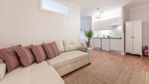 En moderne stue med en beige sofa, mønstrede puder, en lille plante og et køkken med hvide skabe og et hængende lys.
