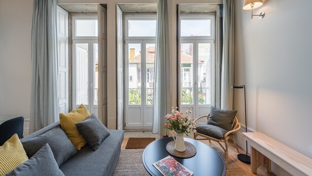A living room with a sofa, armchair, and coffee table, large windows with curtains, and a wall-mounted lamp. - GuestReady - Visconde Mezzanine (Porto)