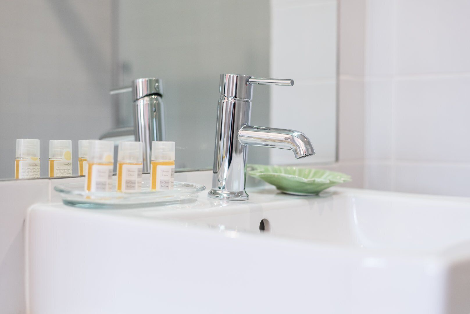 A bathroom sink with two chrome faucets, a glass soap dish, and several small bottles of soap.